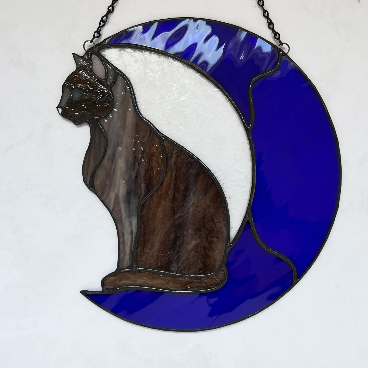 A multi-hued brown and tan stained glass cat sitting on a bright blue moon. This stained glass suncatcher includes a clear leafy looking glass between the cat and the sickle moon. The blue glass is a wavy glass. The eye of the cat is iridescent blue and she has light pink ears. The brown glass is textured like real fur. This picture is on a white background and you can really see the texture of the fur and the ripple of the blue moon glass
