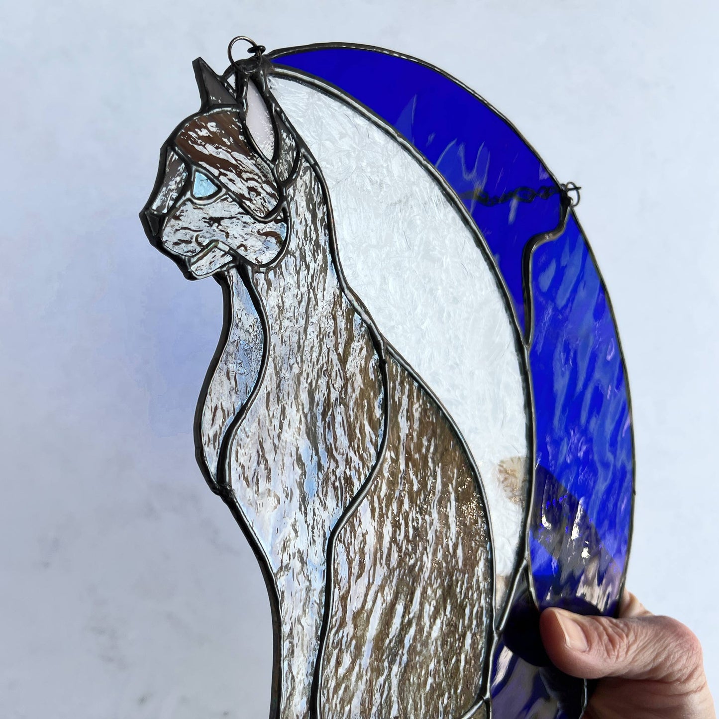 A multi-hued brown and tan stained glass cat sitting on a bright blue moon. This stained glass suncatcher includes a clear leafy looking glass between the cat and the sickle moon. The blue glass is a wavy glass. The eye of the cat is iridescent blue and she has light pink ears. The brown glass is textured like real fur. This picture really shows off the textured fur.