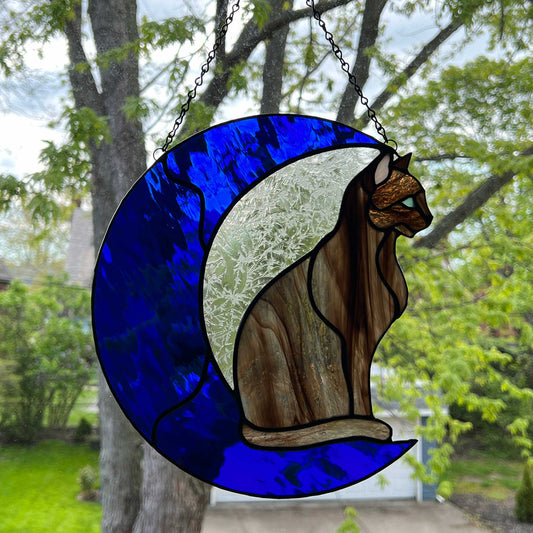 A multi-hued brown and tan stained glass cat sitting on a bright blue moon.  This stained glass suncatcher includes a clear leafy looking glass between the cat and the sickle moon. The blue glass is a wavy glass. The eye of the cat is iridescent blue and she has light pink ears.
