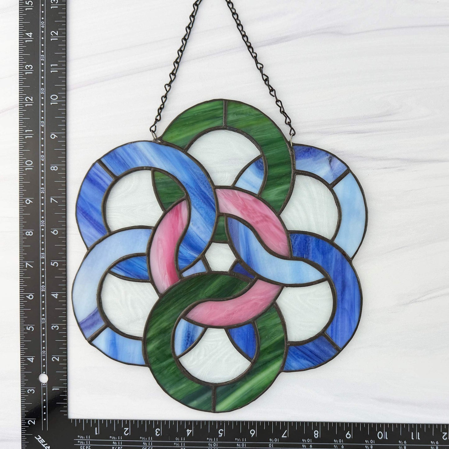 Stained glass circular celtic design with interlocking rings in blue, green, and pink, hanging on a white background with a ruler for scale.
