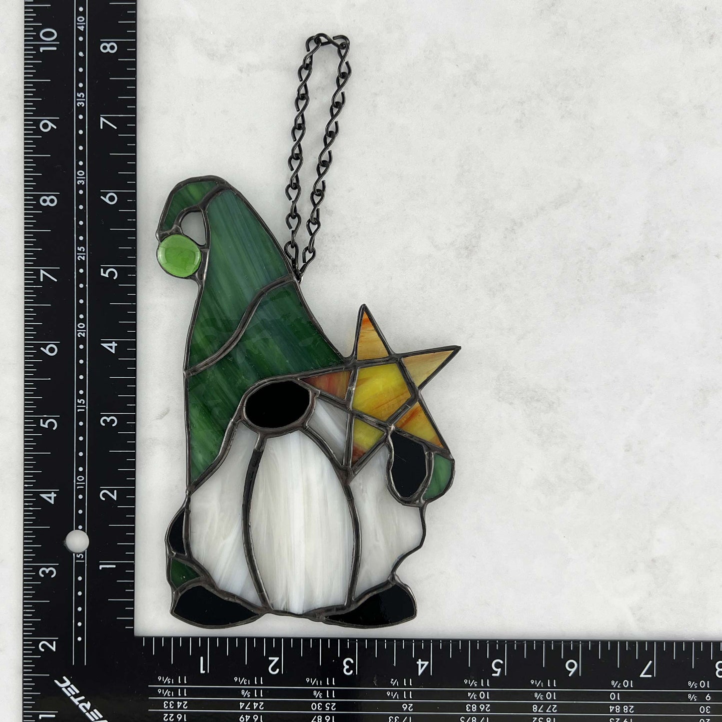 A ruler for sizing of a gnome with a green hat, black mittens and feet and a light green ball at the end of the hat. He's holding a yellow star in his right hand.