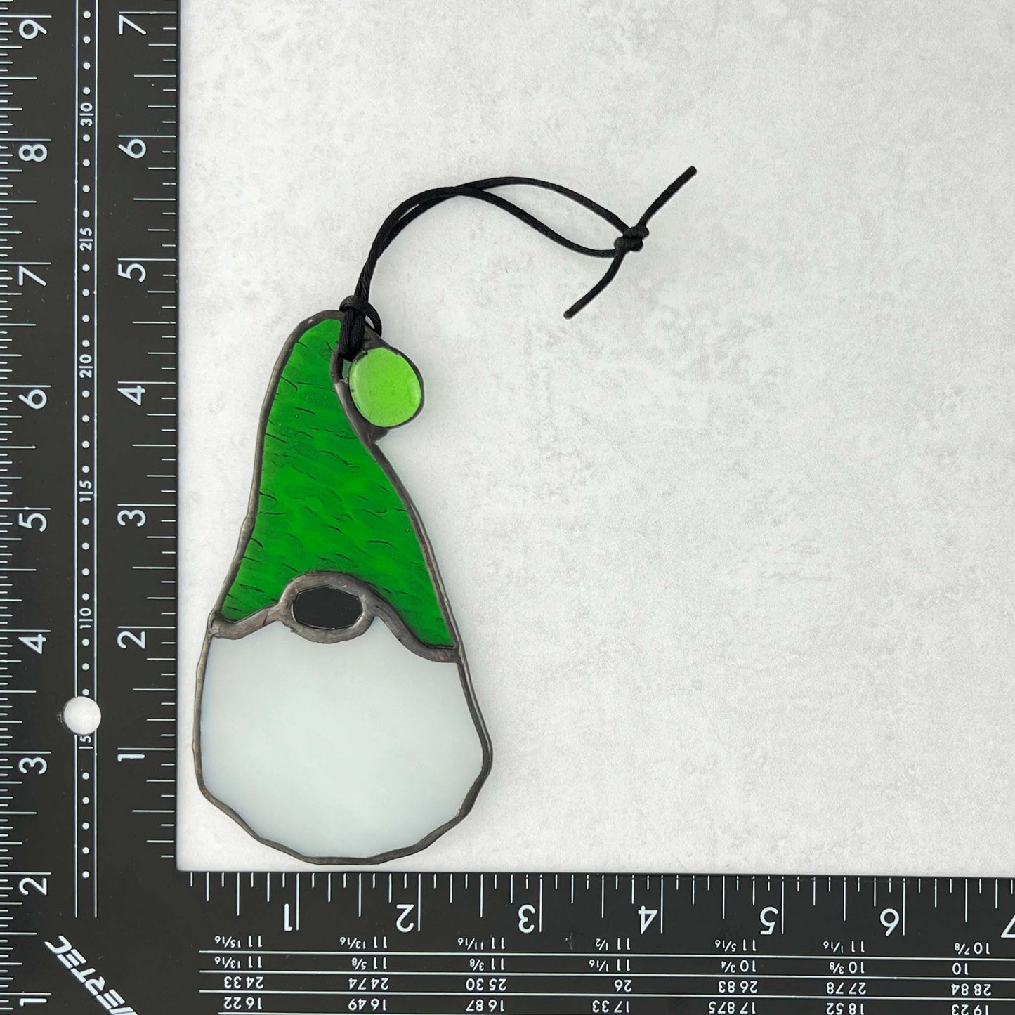 Stained glass gnome ornament with green hat and white beard on a white background with ruler for scale.