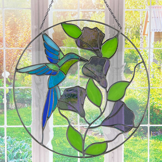 Round stained glass panel with a hummingbird in multiple shades of blue and green sipping out of purple bellflowers shown hanging in a window with a garden background