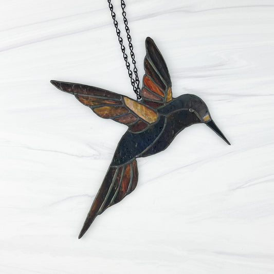 Stained glass hummingbird suncatcher made from textured glass in hues of red, yellow, orange, blue, and earthy brown. The wings are outstretched as if the bird is in midflight.
