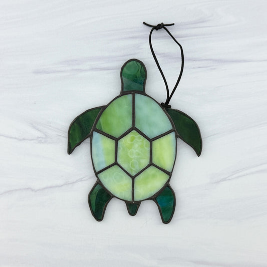 Stained glass turtle suncatcher with a shell of mottled light green opaque glass and flippers and head of translucent blue-green glass. It's displayed on a white background