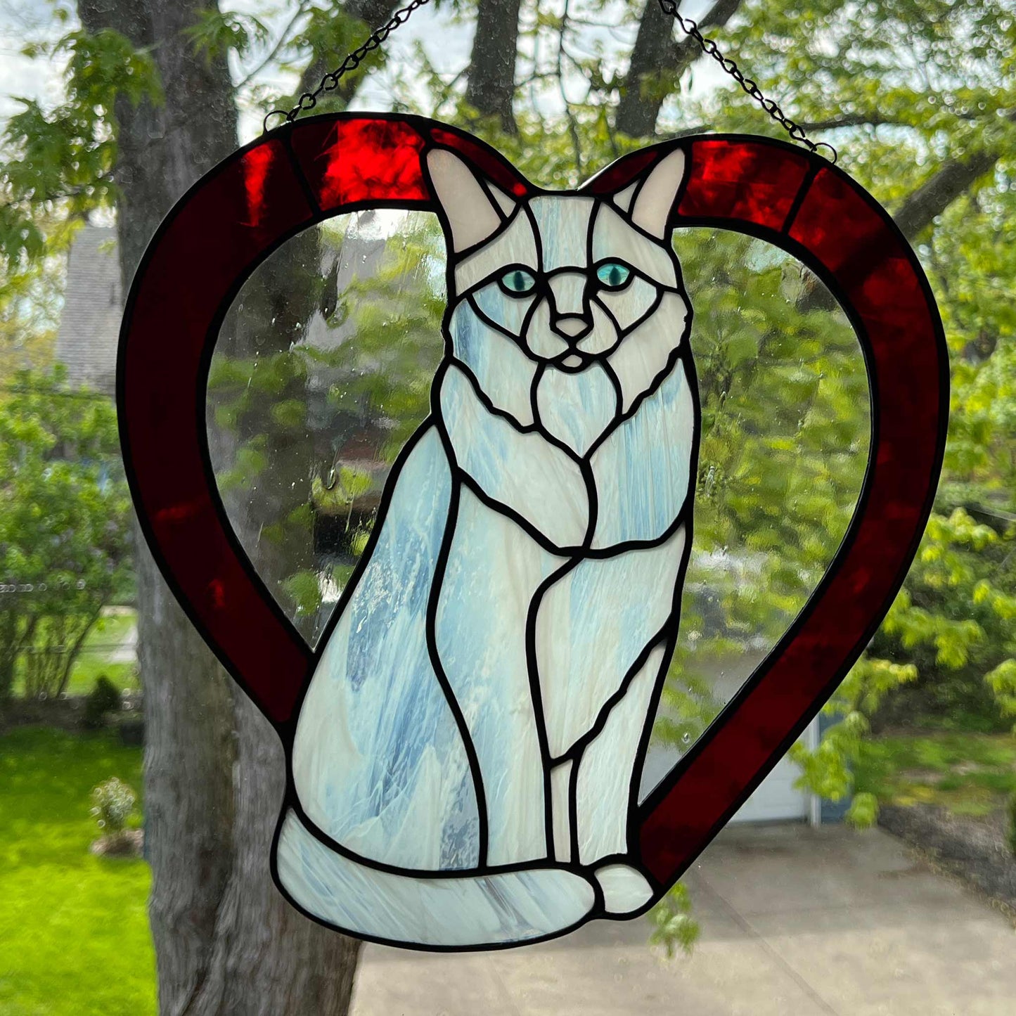 An all white stained glass sitting cat surrounded by a red heart. The glass used for the white fur is textured to resemble real fur and the eyes are an iridescent blue. Light pink is used for the inner ear and the nose. A clear rainy glass is used between the cat and the heart for stability. This picture shows the back side of the cat.