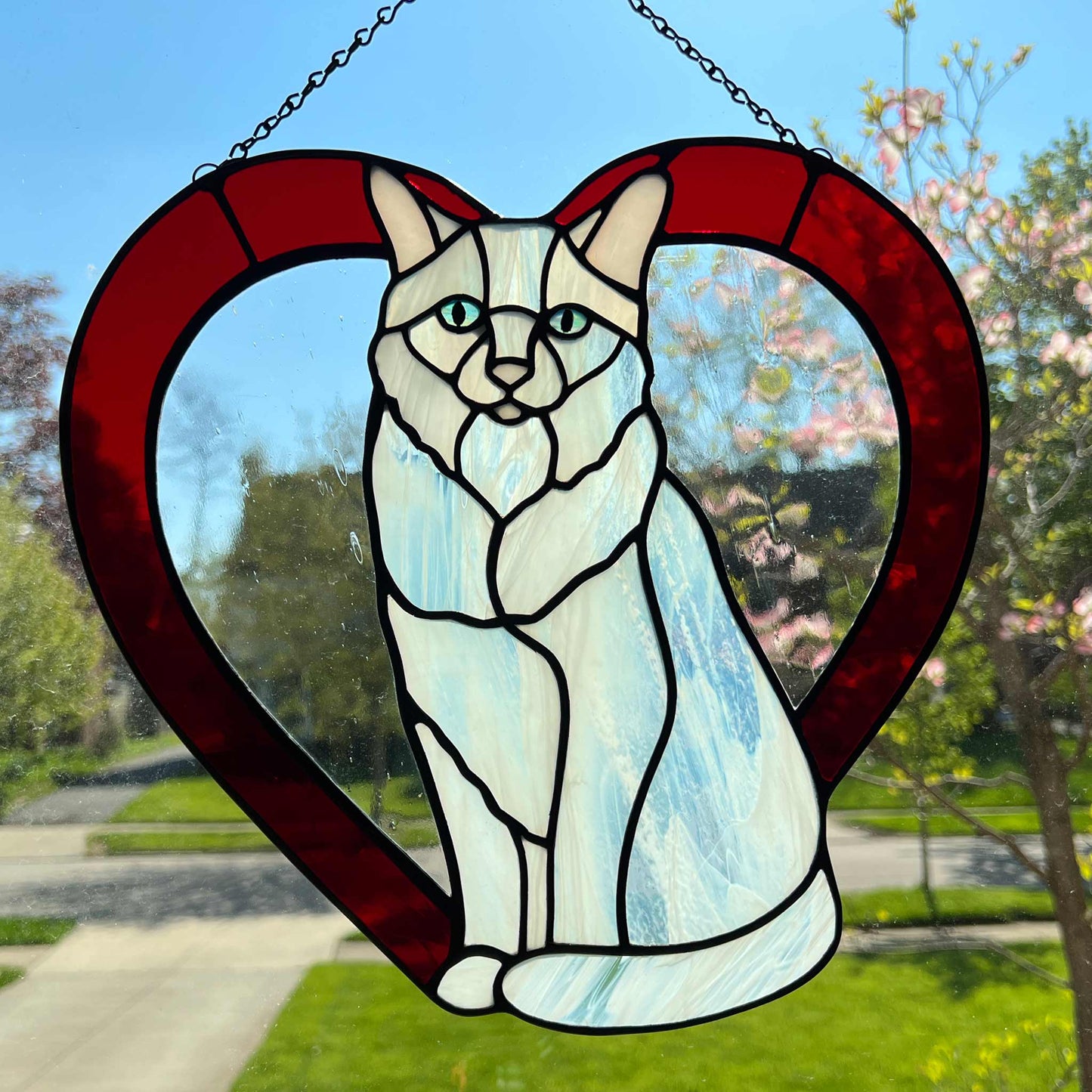 An all white stained glass sitting cat surrounded by a red heart. The glass used for the white fur is textured to resemble real fur and the eyes are an iridescent blue. Light pink is used for the inner ear and the nose. A clear rainy glass is used between the cat and the heart for stability.