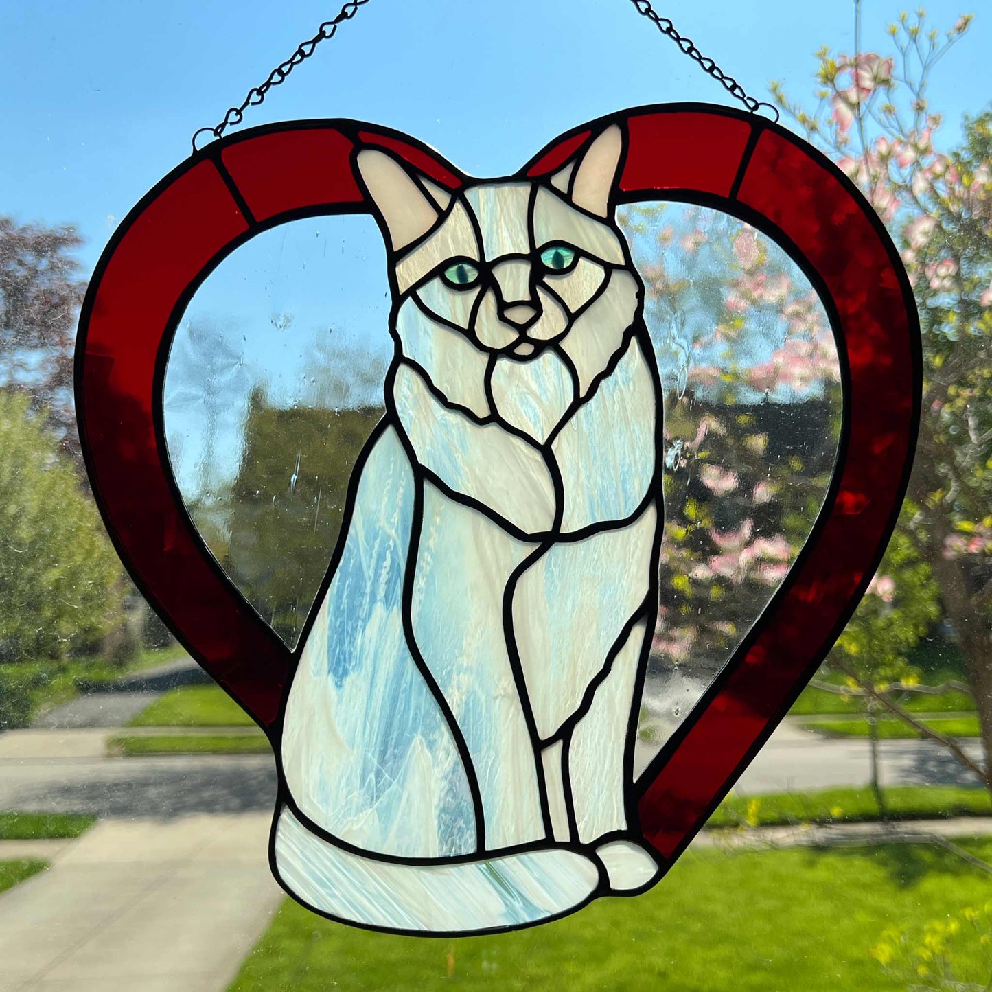 An all white stained glass sitting cat surrounded by a red heart. The glass used for the white fur is textured to resemble real fur and the eyes are an iridescent blue.  Light pink is used for the inner ear and the nose. A clear rainy glass is used between the cat and the heart for stability. This picture is the backside of the cat