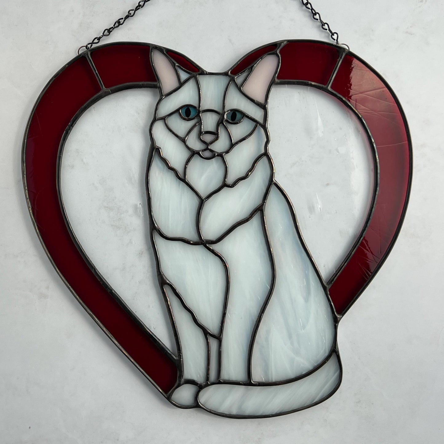 An all white stained glass sitting cat surrounded by a red heart. The glass used for the white fur is textured to resemble real fur and the eyes are an iridescent blue. Light pink is used for the inner ear and the nose. A clear rainy glass is used between the cat and the heart for stability. This pictures shows the cat on a white background and you can see the textured lines in the red glass.