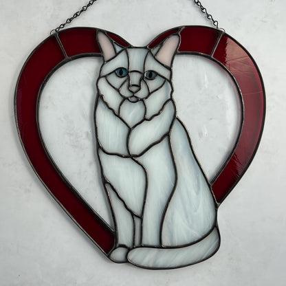 An all white stained glass sitting cat surrounded by a red heart. The glass used for the white fur is textured to resemble real fur and the eyes are an iridescent blue.  Light pink is used for the inner ear and the nose. A clear rainy glass is used between the cat and the heart for stability. This pictures shows the cat on a white background and you can see the textured lines in the red glass.
