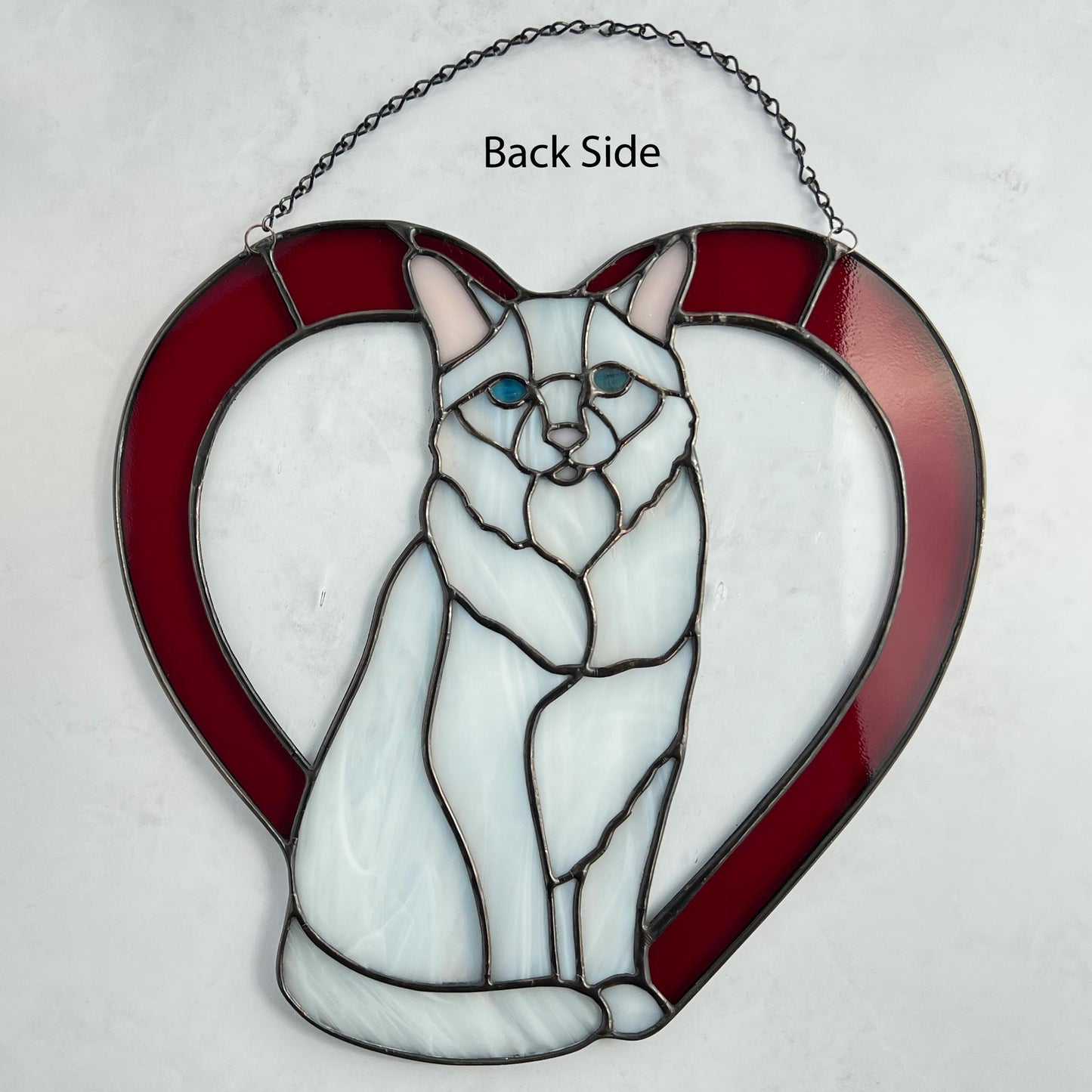 An all white stained glass sitting cat surrounded by a red heart. The glass used for the white fur is textured to resemble real fur and the eyes are an iridescent blue.  Light pink is used for the inner ear and the nose. A clear rainy glass is used between the cat and the heart for stability. This picture shows the back side of the cat on a white background.