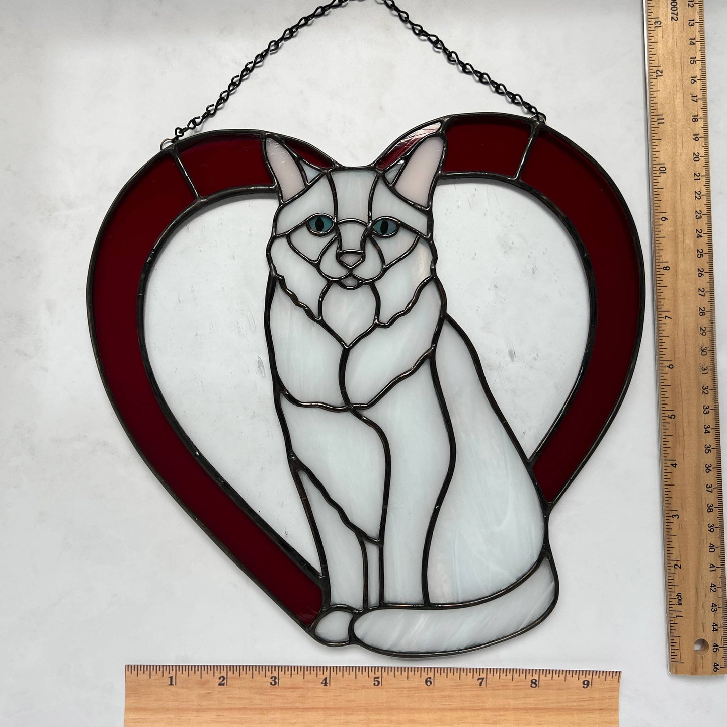 An all white stained glass sitting cat surrounded by a red heart. The glass used for the white fur is textured to resemble real fur and the eyes are an iridescent blue. Light pink is used for the inner ear and the nose. A clear rainy glass is used between the cat and the heart for stability. This picture includes rulers so that you can see that the cat is 11.25 inches wide by 11 inches tall.
