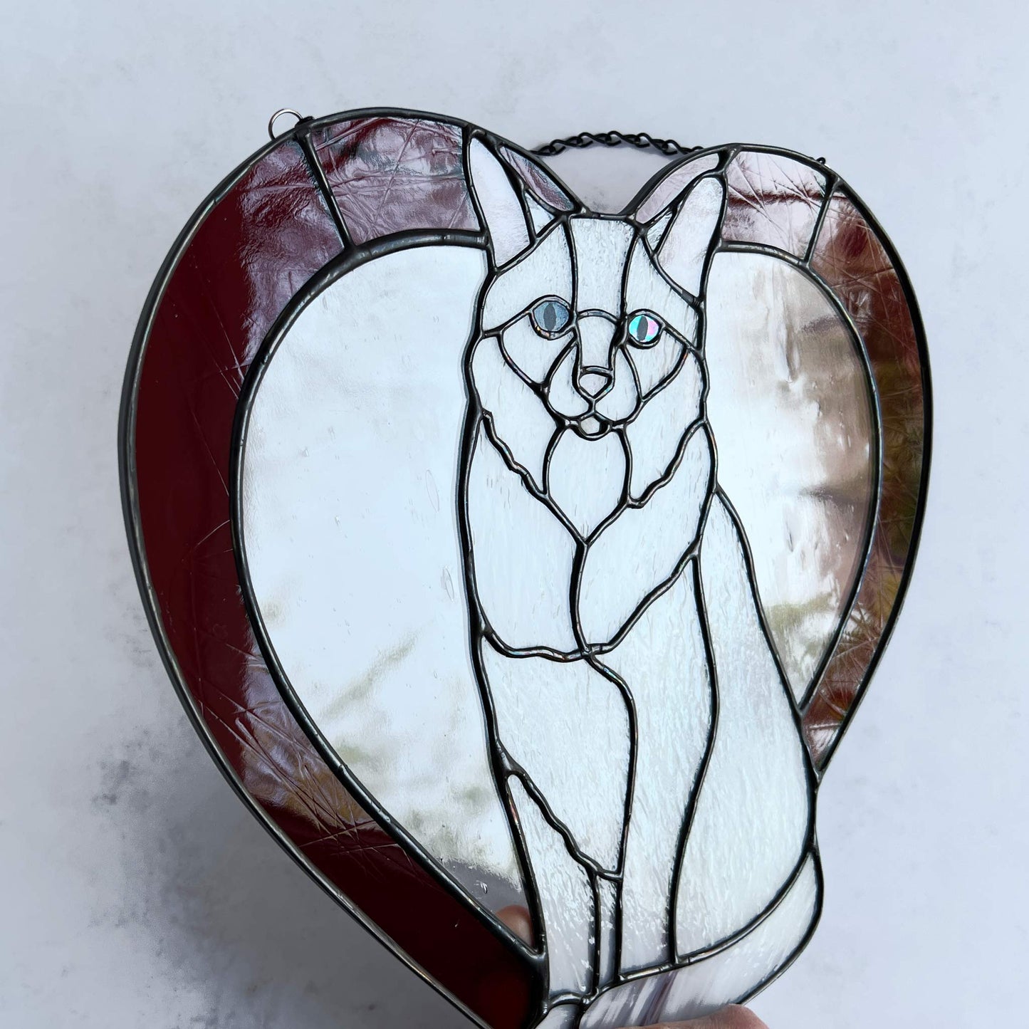 An all white stained glass sitting cat surrounded by a red heart. The glass used for the white fur is textured to resemble real fur and the eyes are an iridescent blue. Light pink is used for the inner ear and the nose. A clear rainy glass is used between the cat and the heart for stability. This picture shows the texture of the red glass and you can see the iridescence of the eye