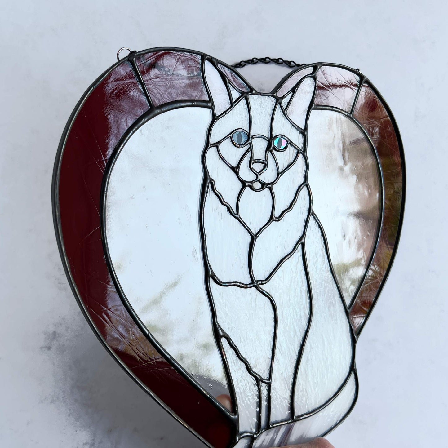 An all white stained glass sitting cat surrounded by a red heart. The glass used for the white fur is textured to resemble real fur and the eyes are an iridescent blue.  Light pink is used for the inner ear and the nose. A clear rainy glass is used between the cat and the heart for stability. This picture shows the texture of the red glass and you can see the iridescence of the eye