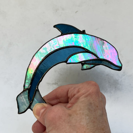 This dolphin suncatcher captures the grace and playfulness of one of the ocean's most beloved creatures. This suncatcher features a light blue iridescent glass for the majority of the dolphin's body, with a darker blue glass used for the belly and top fin. It measures 5.5x4.5 inches and will appeal to animal lovers!