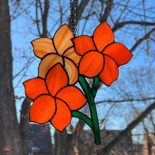 Stained glass suncatcher features three intricate Frangipani flowers in various shades of orange, perfectly capturing the delicate beauty of this tropical blossom with green stems. It measures 7x6.5 inches.