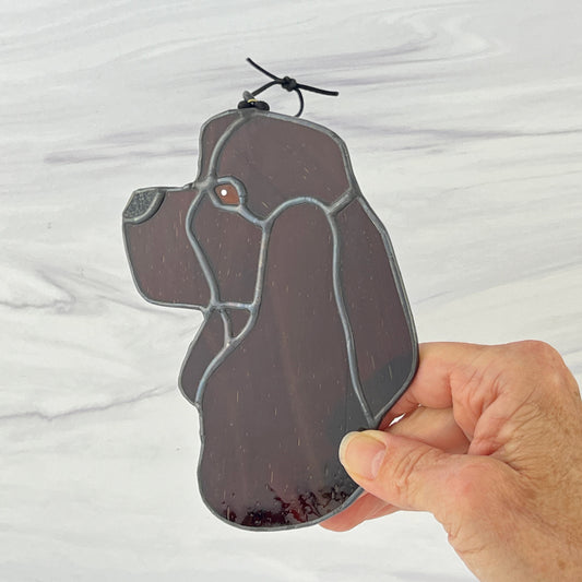 A hand holding the stained glass Chocolate American Cocker Spaniel dog head suncatcher.  The varied brown shades are clearly visible in this view and the textured nose as well.
