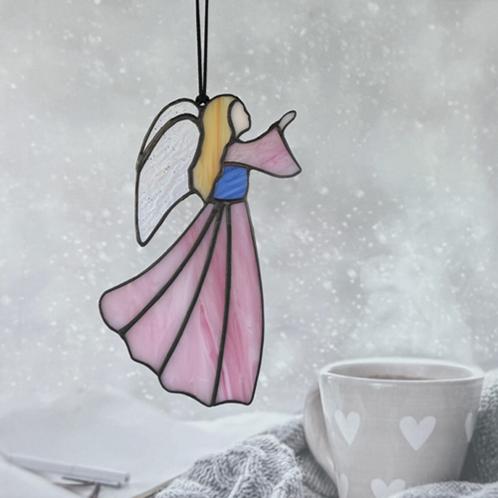 Pink stained glass angel hanging in a window above a coffee mug with a snowy background. The angel has her hand raised to the right. She has long blonde hair, irridescent wings and her bodice is blue.