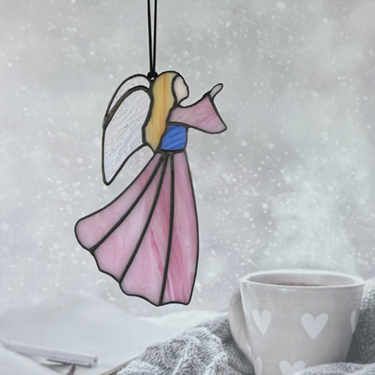 Pink stained glass angel hanging in a window above a coffee mug with a snowy background. The angel has her hand raised to the right. She has long blonde hair, irridescent wings and her bodice is blue.
