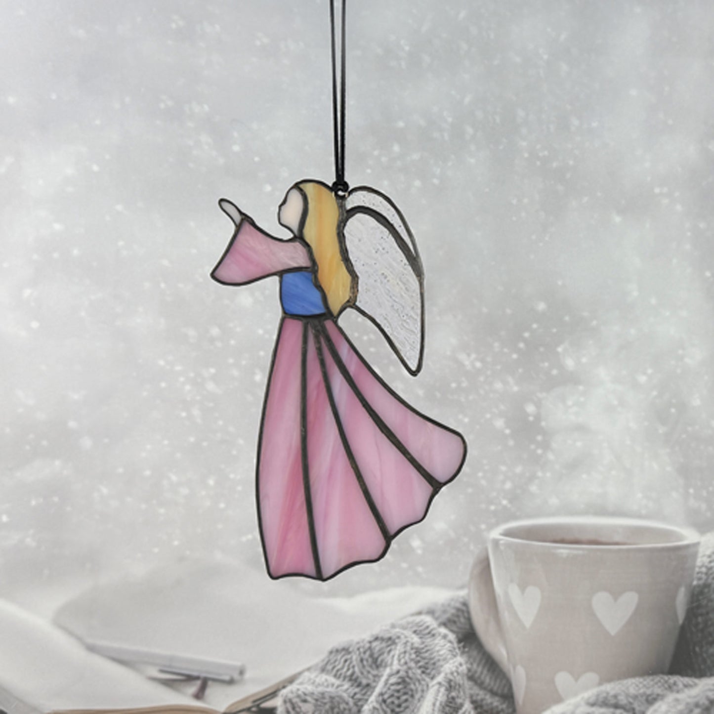 Pink stained glass angel hanging on a black cord with a cozy indoor background. Her hair is golden, her bodice blue and she is looking to the left.
