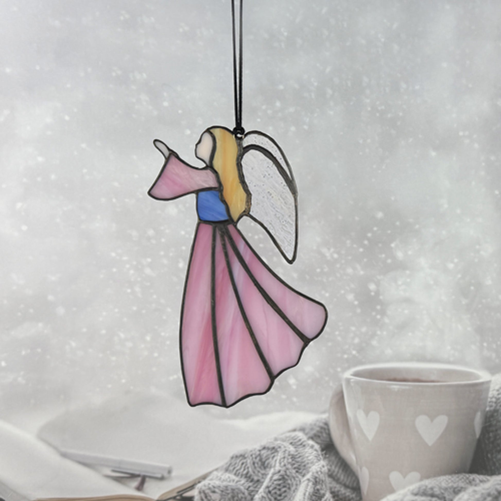 Pink stained glass angel hanging on a black cord with a cozy indoor background. Her hair is golden, her bodice blue and she is looking to the left.