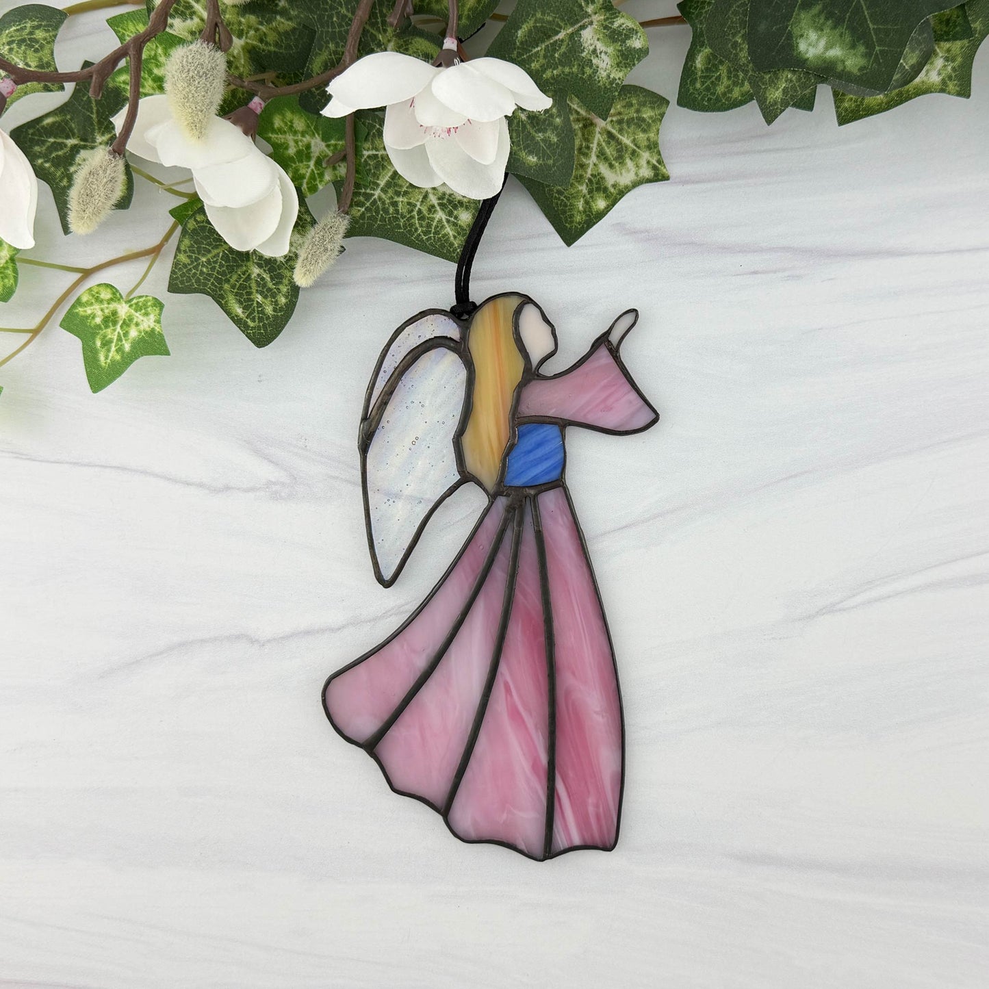 Stained glass angel decoration with pink wings and blonde hair, placed on a white surface with green leaves and white flowers in the background.
