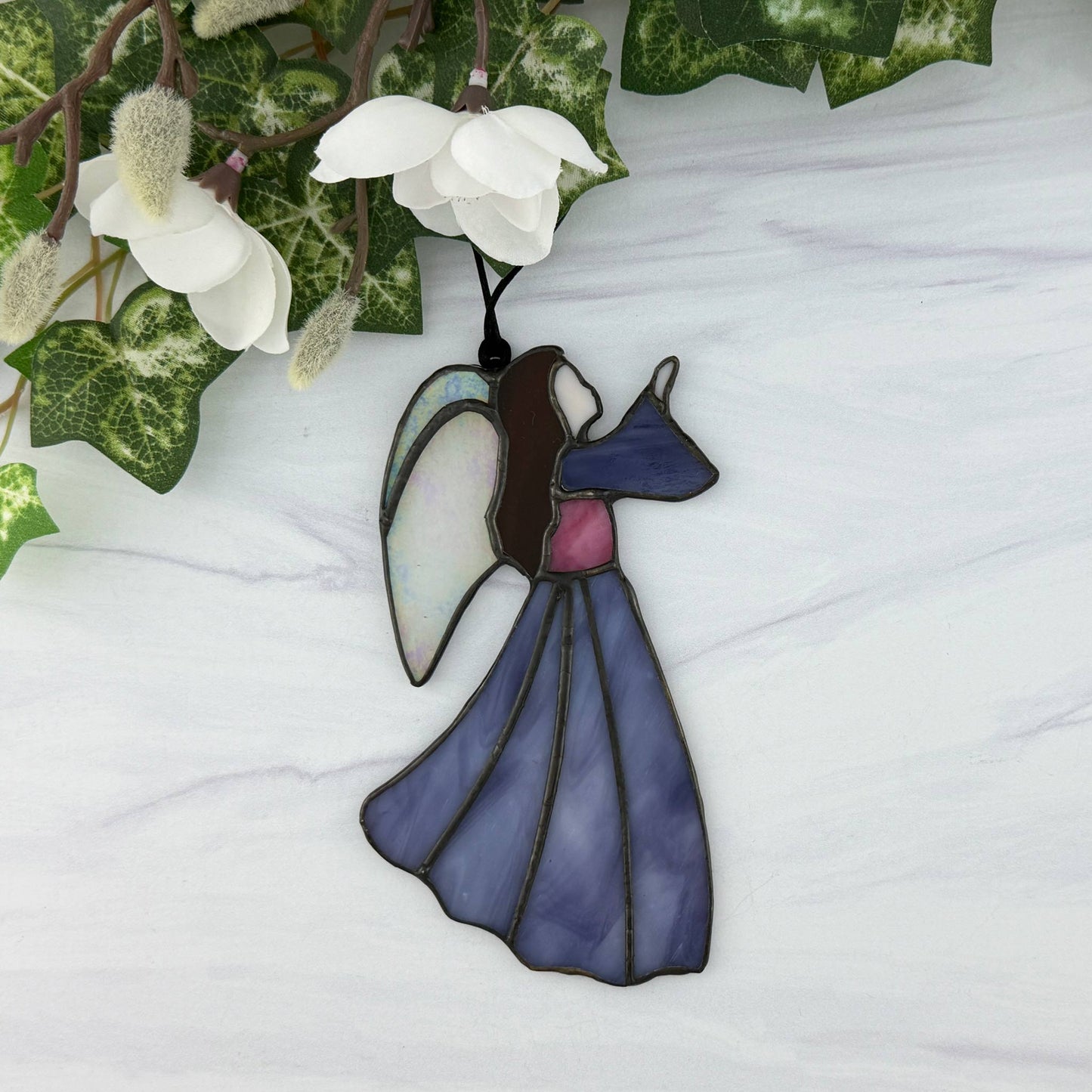 Stained glass purple angel ornament on a white surface with green leaves and white flowers in the background. Her long hair is brown, her bodice is purple and she has irridescent wings.