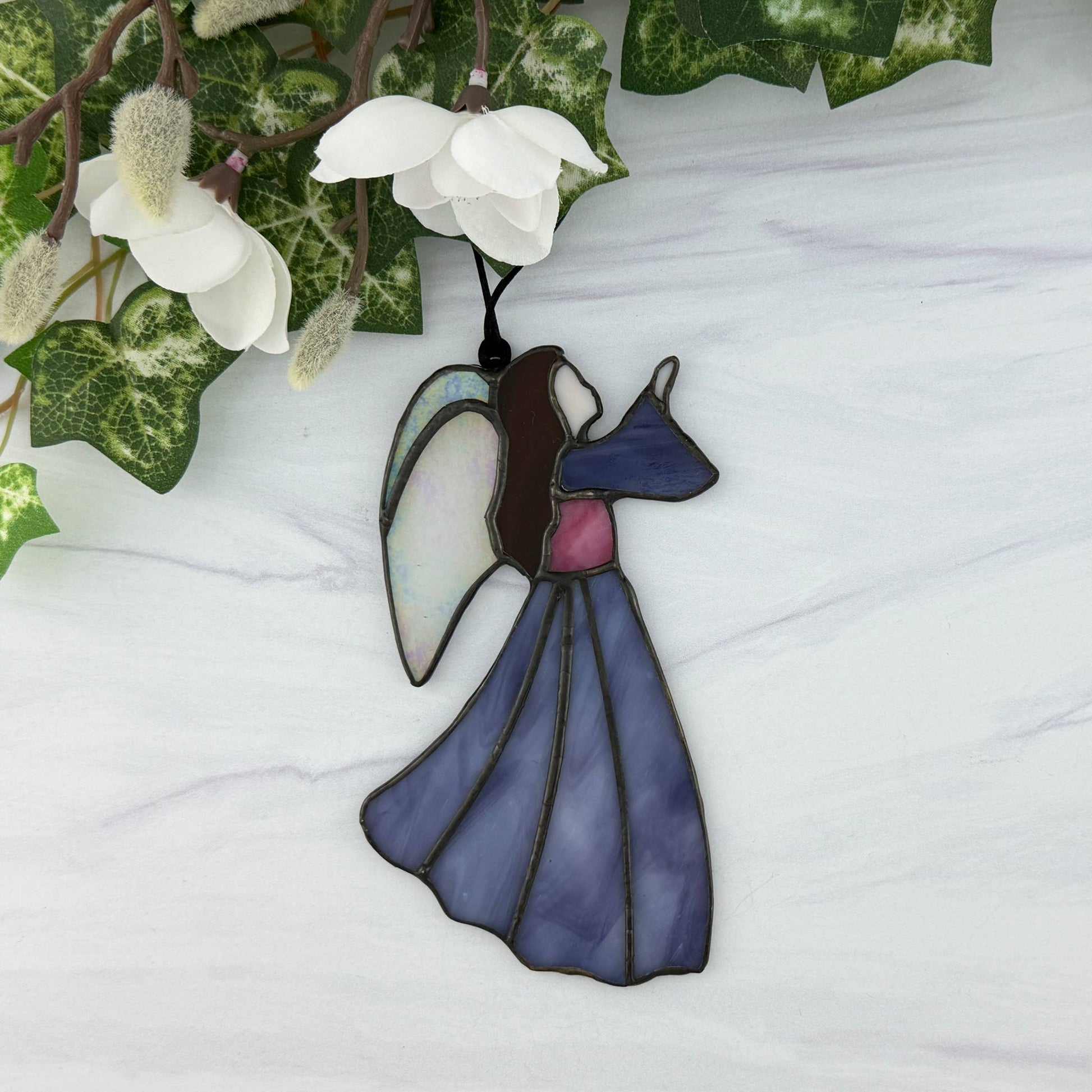 Stained glass purple angel ornament on a white surface with green leaves and white flowers in the background. Her long hair is brown, her bodice is purple and she has irridescent wings.