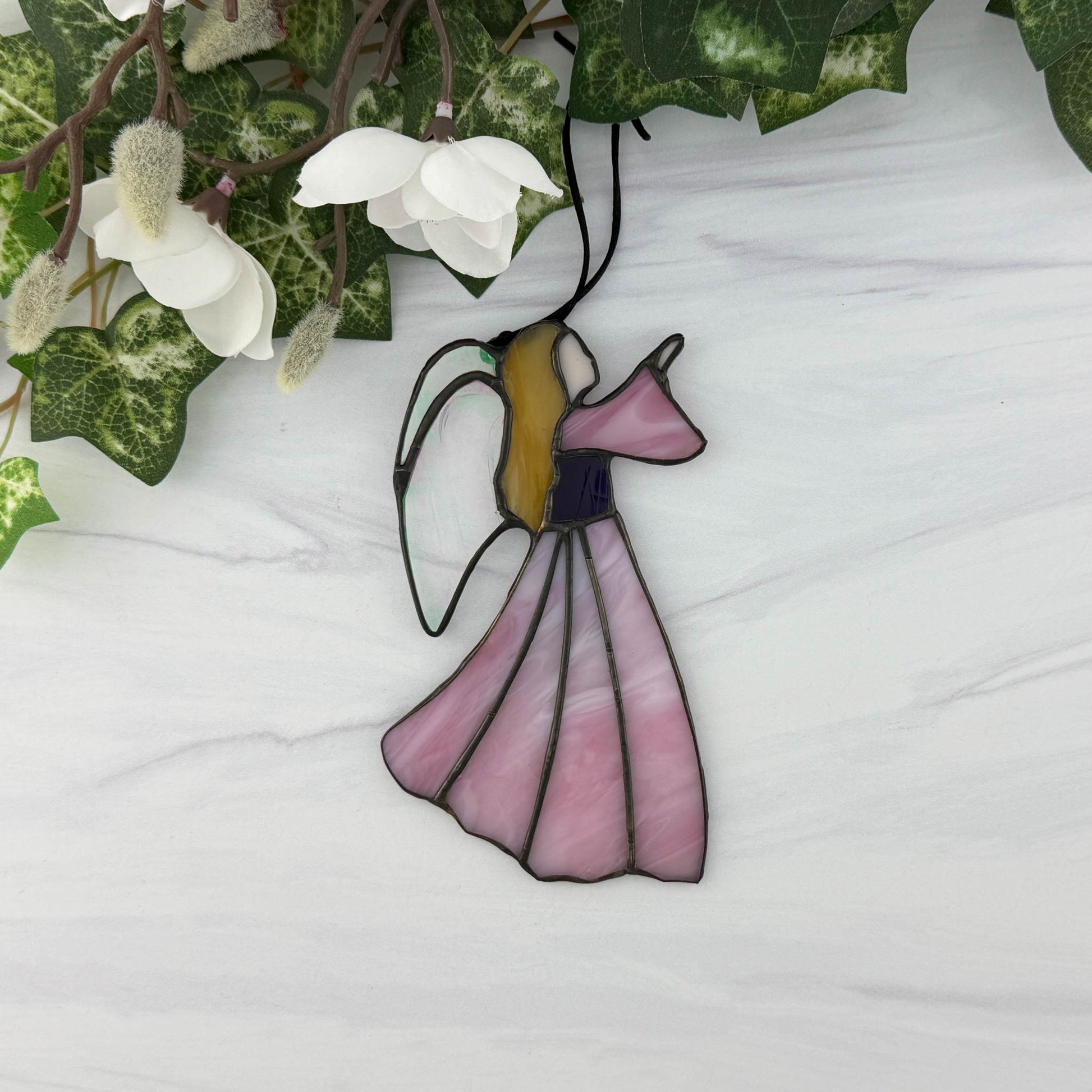 Stained glass angel with a pink dress and clear irridescent wings on a white background with green ivy and white flowers.
