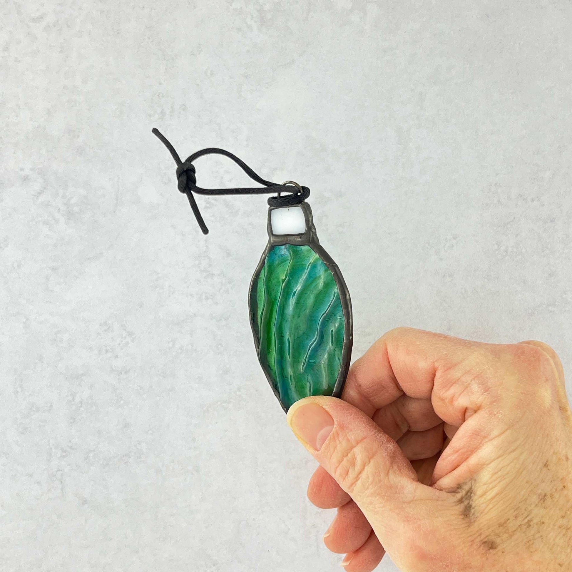 Hand holding a Green-Blue rippled stained glass bulb shaped Christmas ornament hung from a black cord.