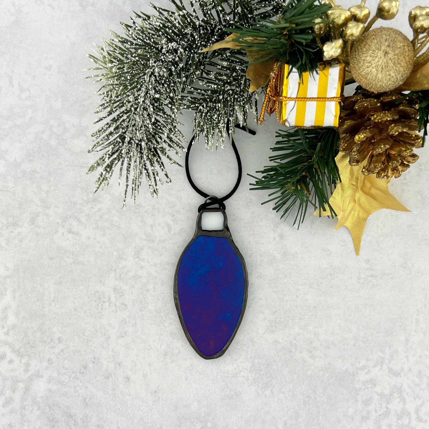 Iridescent Teal-Blue stained glass bulb shaped Christmas ornament hung from a black cord. Shown hanging from a tree branch.
