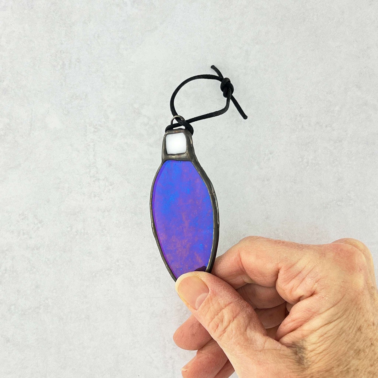 Hand holding an Iridescent Teal-Blue stained glass bulb shaped Christmas ornament hung from a black cord.