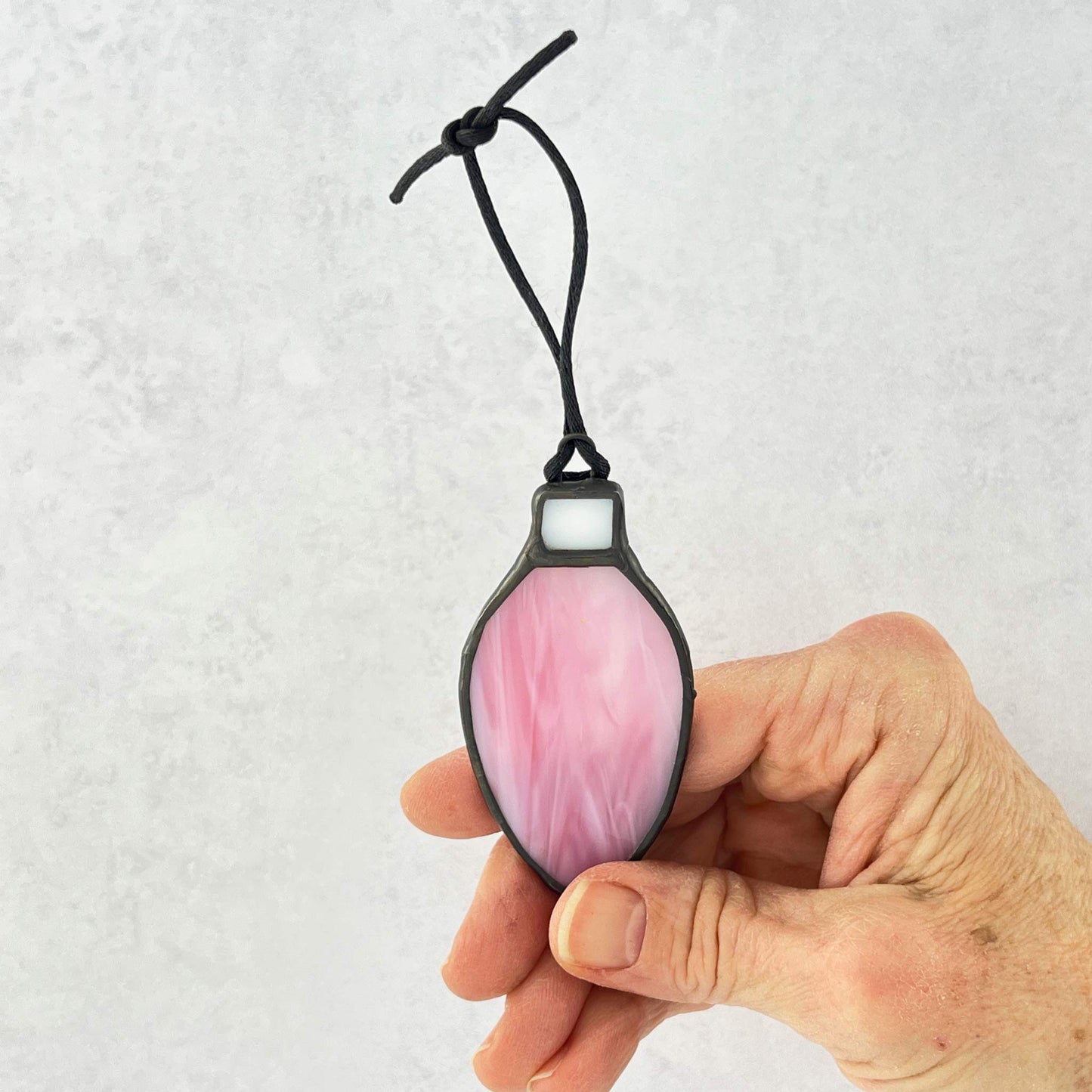 hand holding a pink and white stained glass bulb shaped Christmas ornament hung from a black cord.  