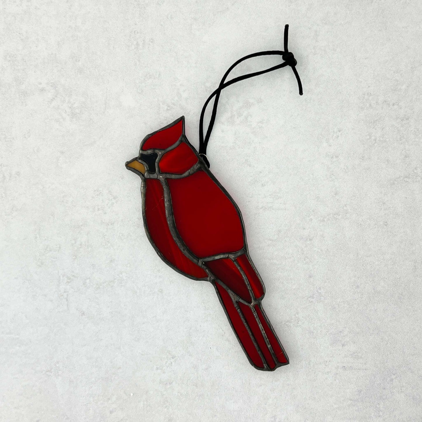 Red stained glass cardinal bird ornament on a white background. Various colors of red glass are used with an orange beak and a black cord for hanging.