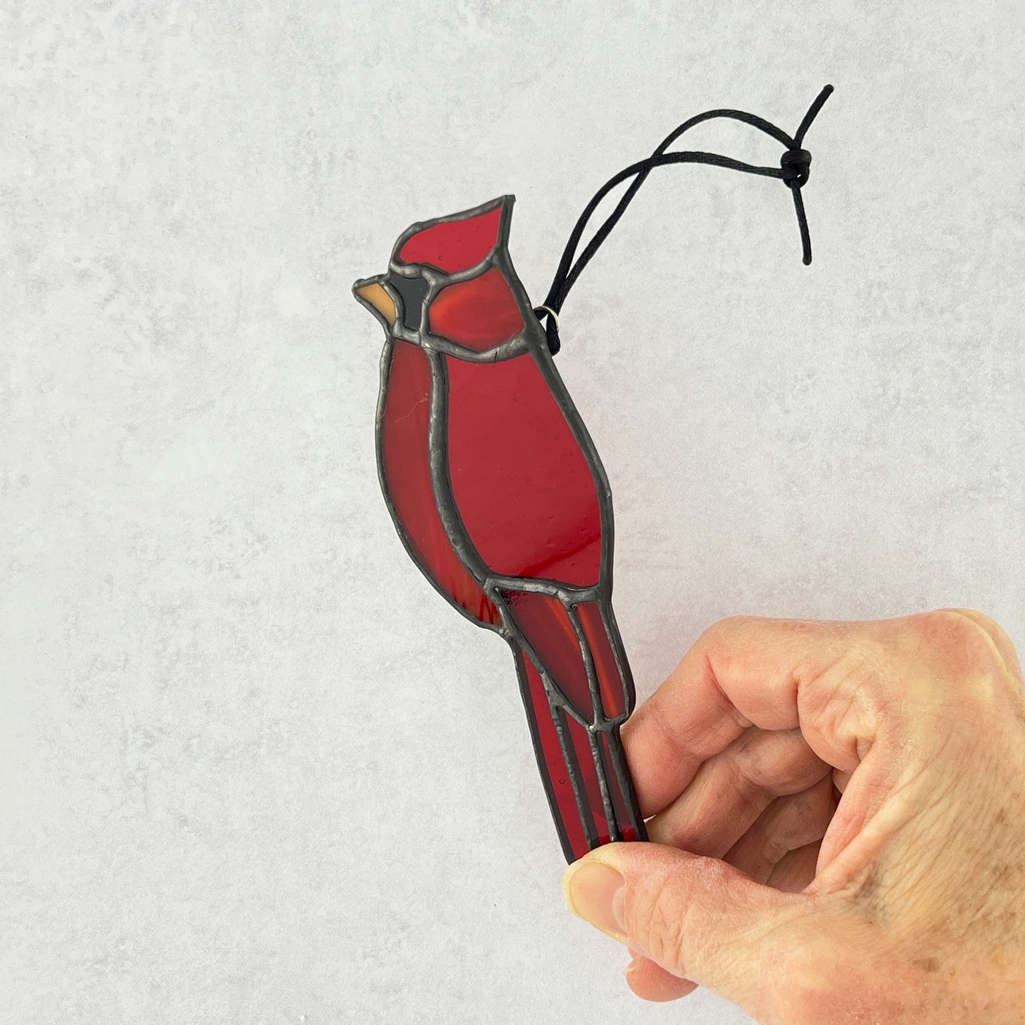 Red stained glass male cardinal bird ornament held by a hand.  Various colors of red glass are used with an orange beak and a black cord for hanging.