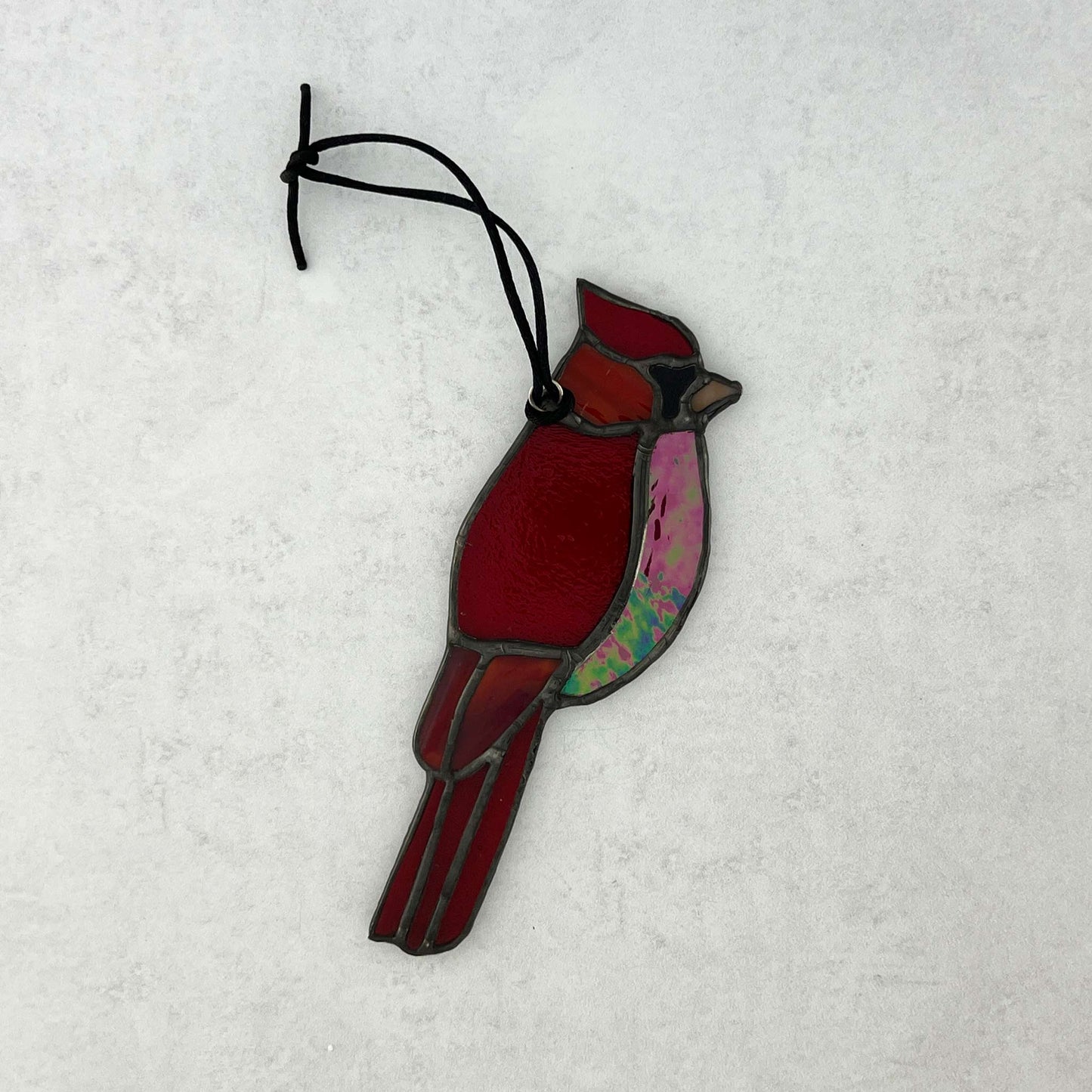 Red stained glass cardinal bird ornament on a white background. Various colors of red glass including an iridescent red glass for the breast and an orange beak. A black cord for hanging is attached.
