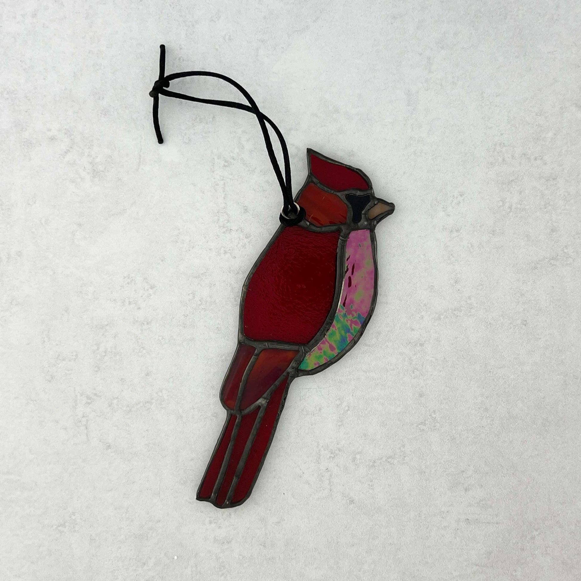 Red stained glass cardinal bird ornament on a white background. Various colors of red glass including an iridescent red glass for the breast and an orange beak. A black cord for hanging is attached.