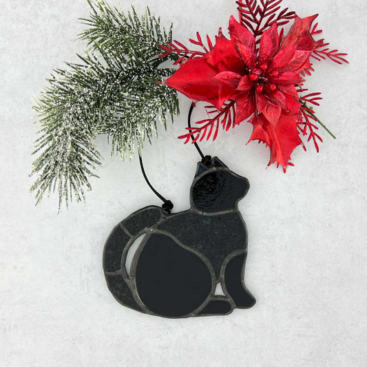 Stained glass Black cat ornament made of smooth and textured black glass. It's displayed with a red poinsettia and greenery on a snowy background