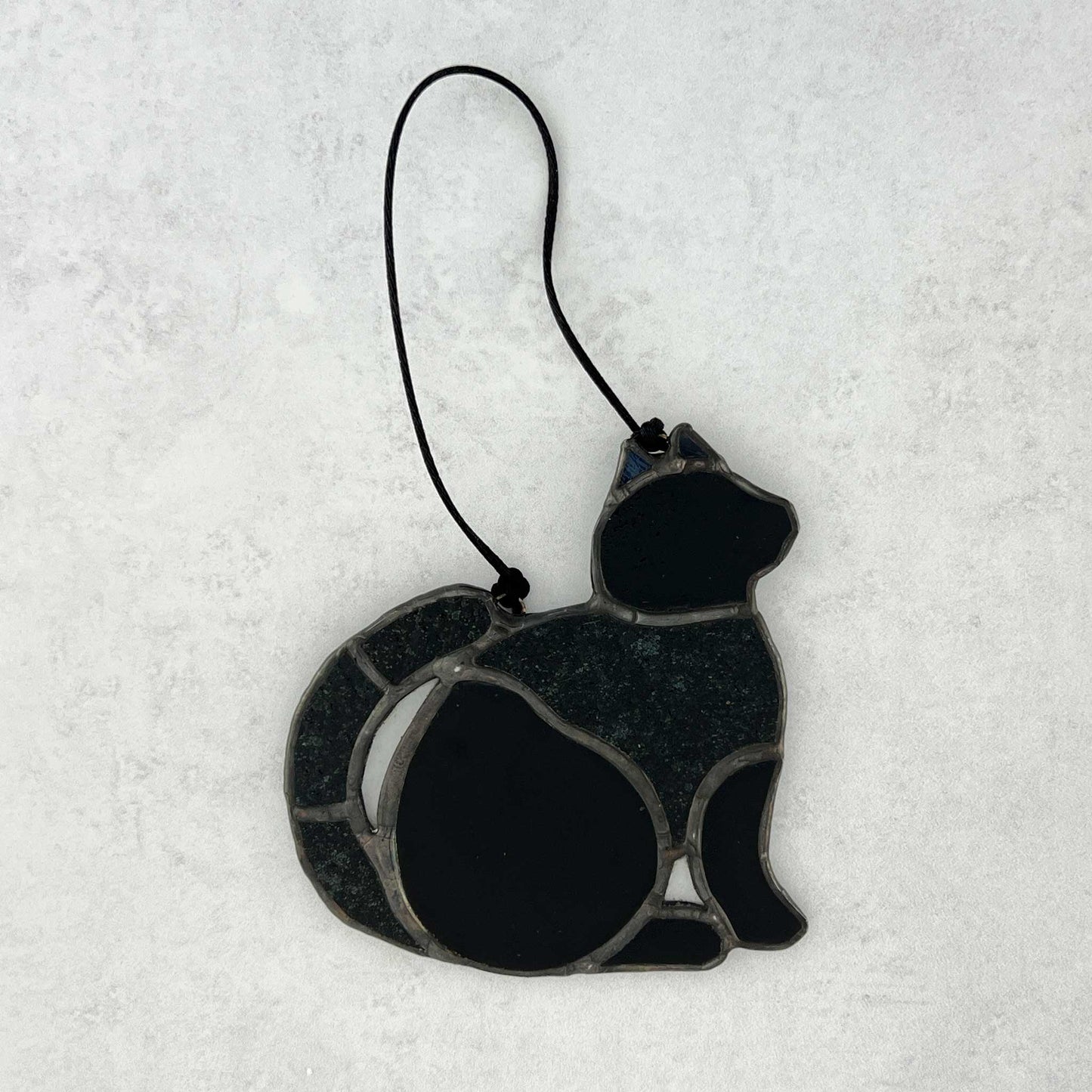 Stained glass Black cat ornament made of smooth and textured black glass. Black cord for hanging is shown.