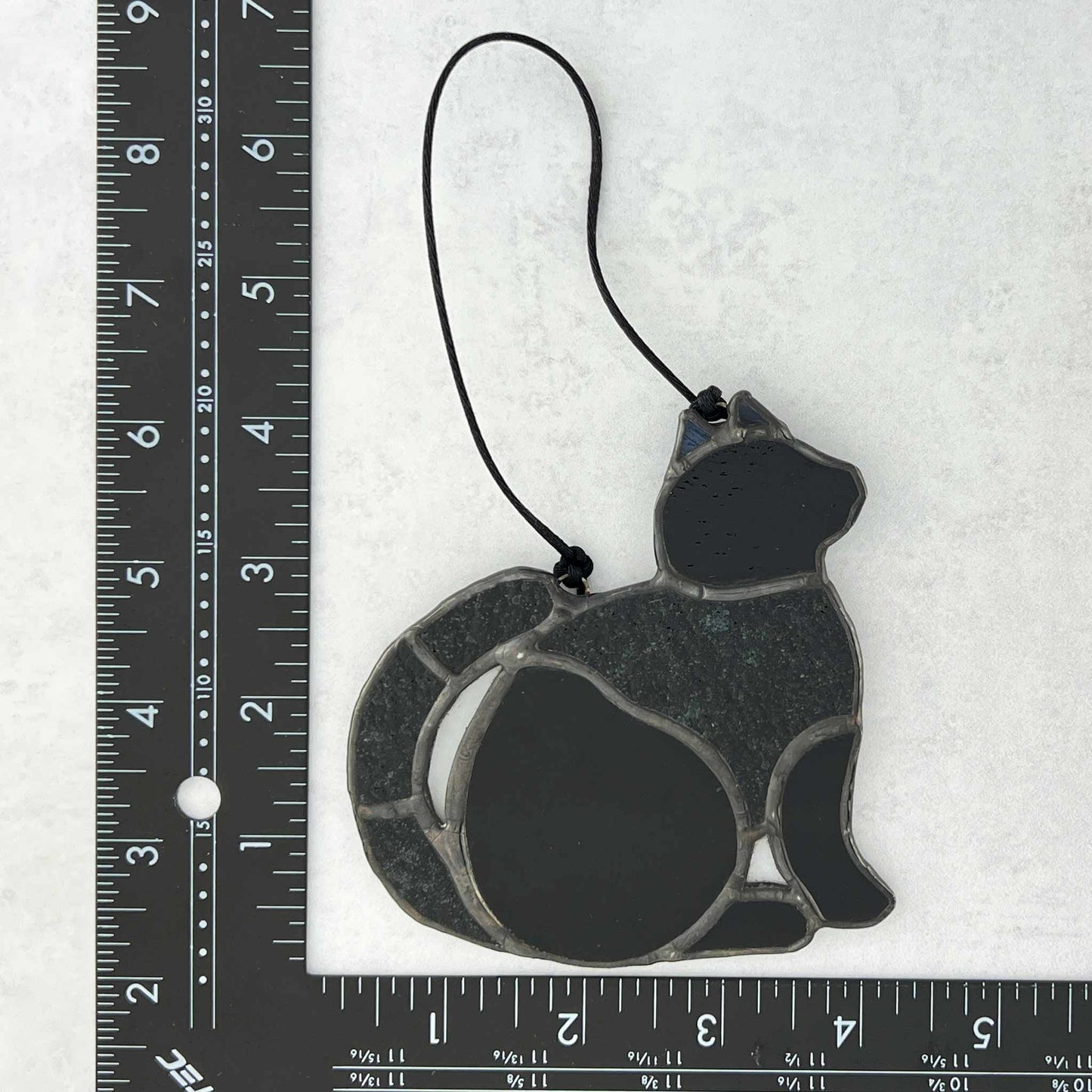 A ruler showing the dimensions of a stained glass Black cat ornament made of smooth and textured black glass. 