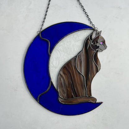 A multi-hued brown and tan stained glass cat sitting on a bright blue moon.  This stained glass suncatcher includes a clear leafy looking glass between the cat and the sickle moon. The blue glass is a wavy glass. The eye of the cat is iridescent blue and she has light pink ears. The brown glass is textured like real fur. This picture shows the iridescent eye better