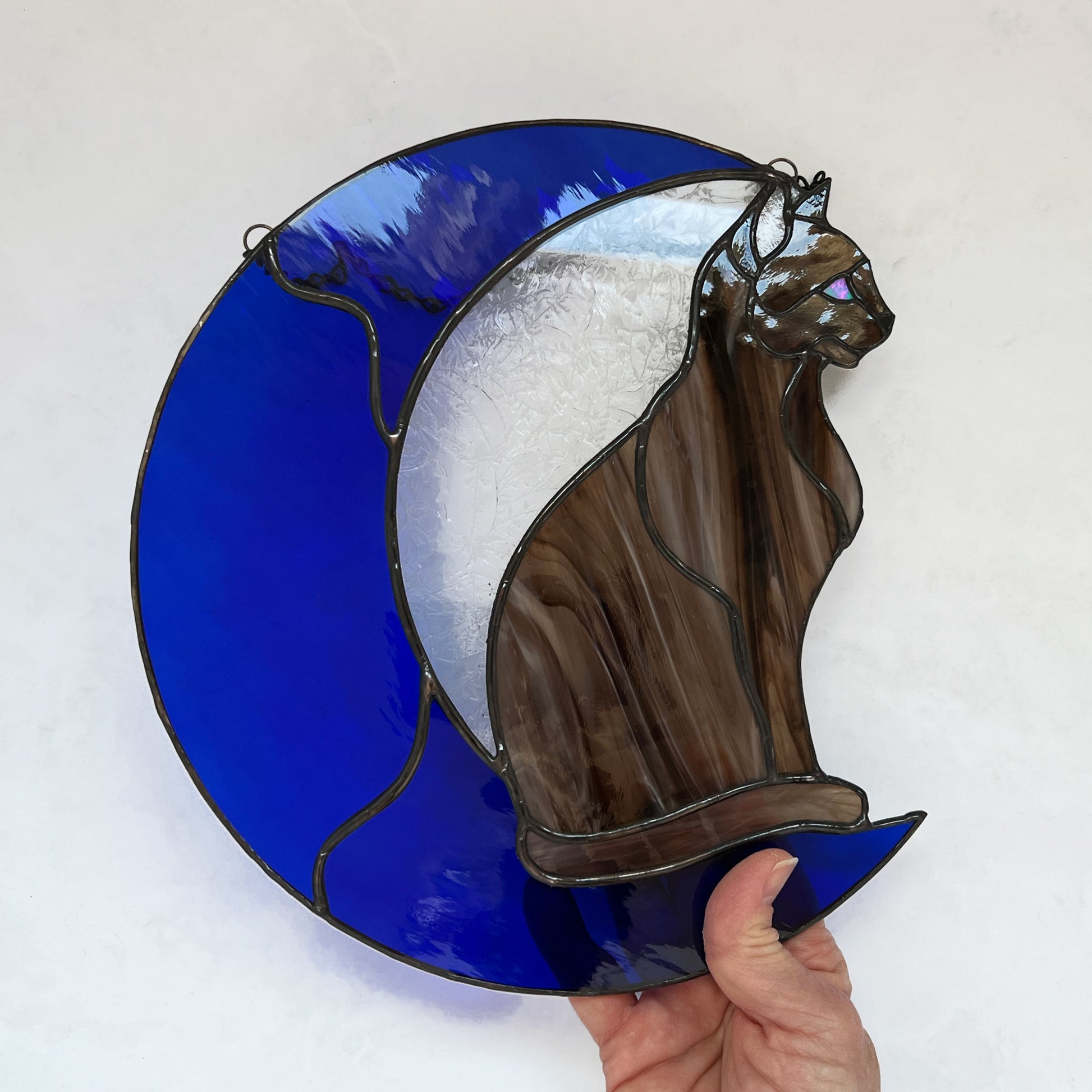 A multi-hued brown and tan stained glass cat sitting on a bright blue moon.  This stained glass suncatcher includes a clear leafy looking glass between the cat and the sickle moon. The blue glass is a wavy glass. The eye of the cat is iridescent blue and she has light pink ears. The brown glass is textured like real fur. A hand is holding the cat in this picture.