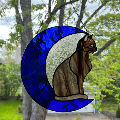 A multi-hued brown and tan stained glass cat sitting on a bright blue moon.  This stained glass suncatcher includes a clear leafy looking glass between the cat and the sickle moon. The blue glass is a wavy glass. The eye of the cat is iridescent blue and she has light pink ears.