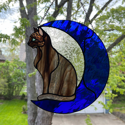 A multi-hued brown and tan stained glass cat sitting on a bright blue moon.  This stained glass suncatcher includes a clear leafy looking glass between the cat and the sickle moon. The blue glass is a wavy glass. The eye of the cat is iridescent blue and she has light pink ears. The brown glass is textured like real fur.