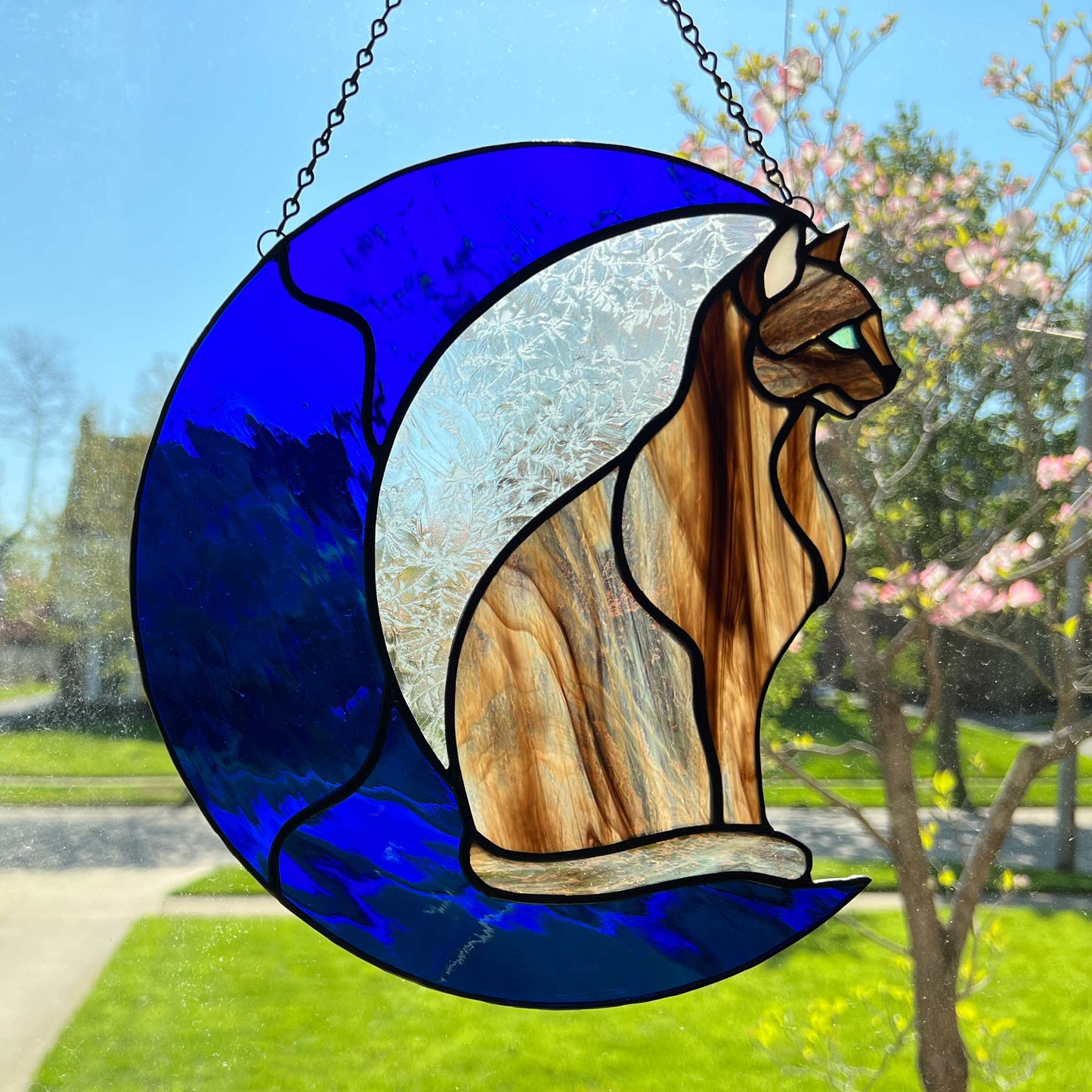 A multi-hued brown and tan stained glass cat sitting on a bright blue moon.  This stained glass suncatcher includes a clear leafy looking glass between the cat and the sickle moon. The blue glass is a wavy glass. The eye of the cat is iridescent blue and she has light pink ears. The brown glass is textured like real fur. This picture is in the bright sun and shows off the variety in the brown fur.