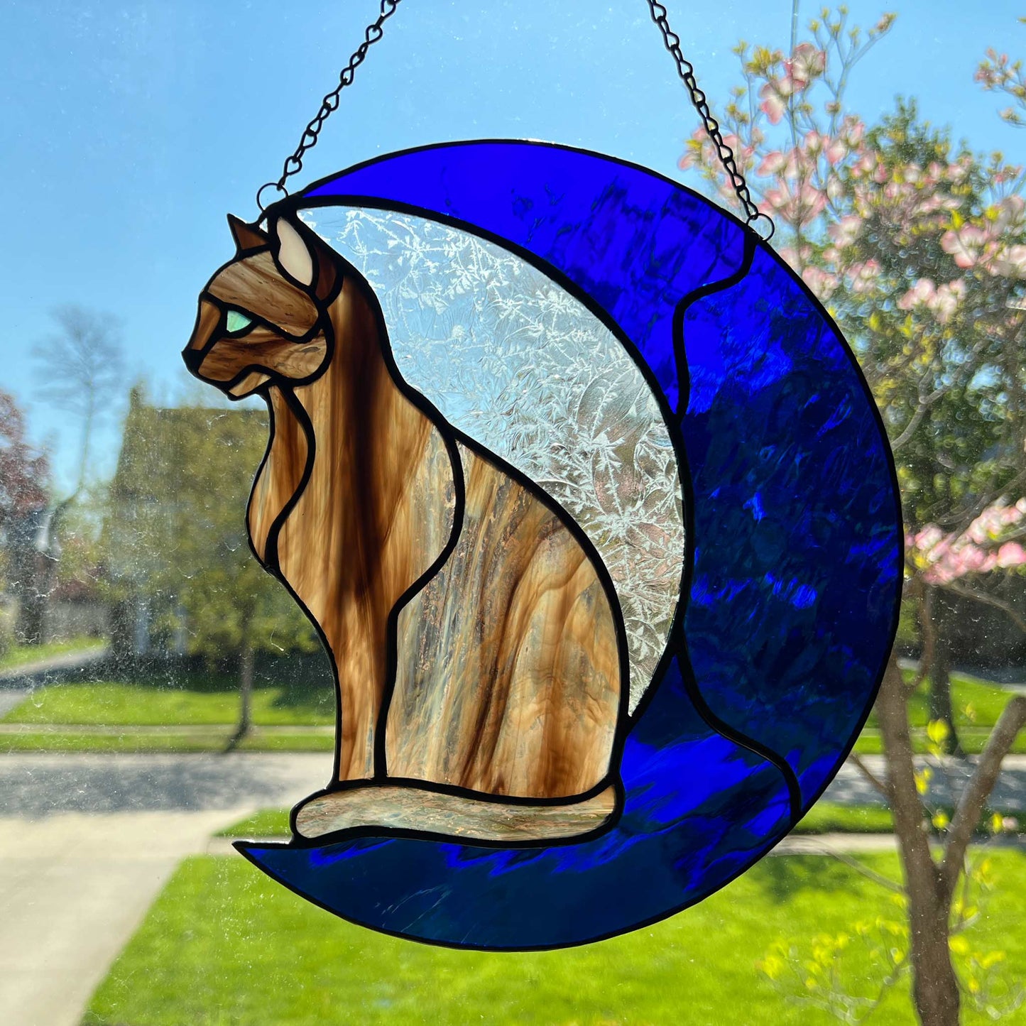 A multi-hued brown and tan stained glass cat sitting on a bright blue moon.  This stained glass suncatcher includes a clear leafy looking glass between the cat and the sickle moon. The blue glass is a wavy glass. The eye of the cat is iridescent blue and she has light pink ears. The brown glass is textured like real fur. This is another picture with the cat facing in the opposite direction.