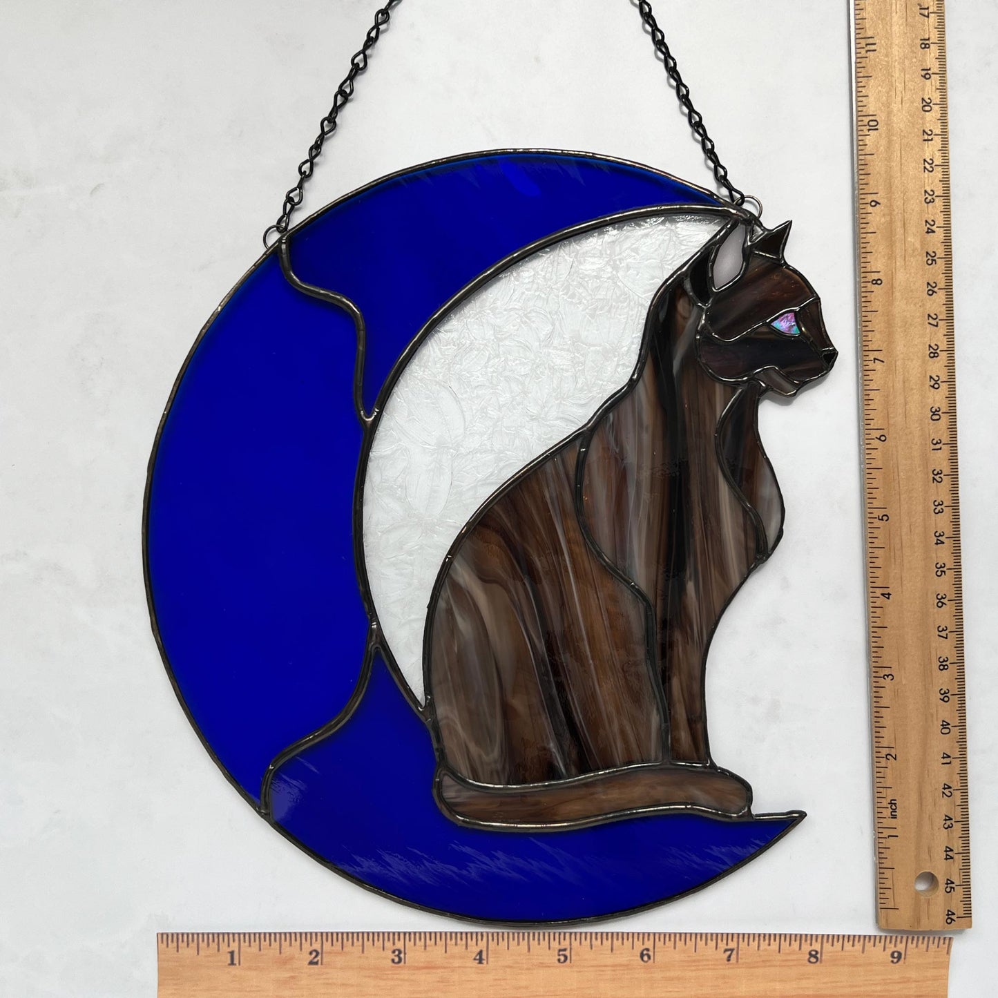 A multi-hued brown and tan stained glass cat sitting on a bright blue moon.  This stained glass suncatcher includes a clear leafy looking glass between the cat and the sickle moon. The blue glass is a wavy glass. The eye of the cat is iridescent blue and she has light pink ears. The brown glass is textured like real fur. Rulers show the size of the suncatcher to be 8 inches wide by 9.75 inches tall.