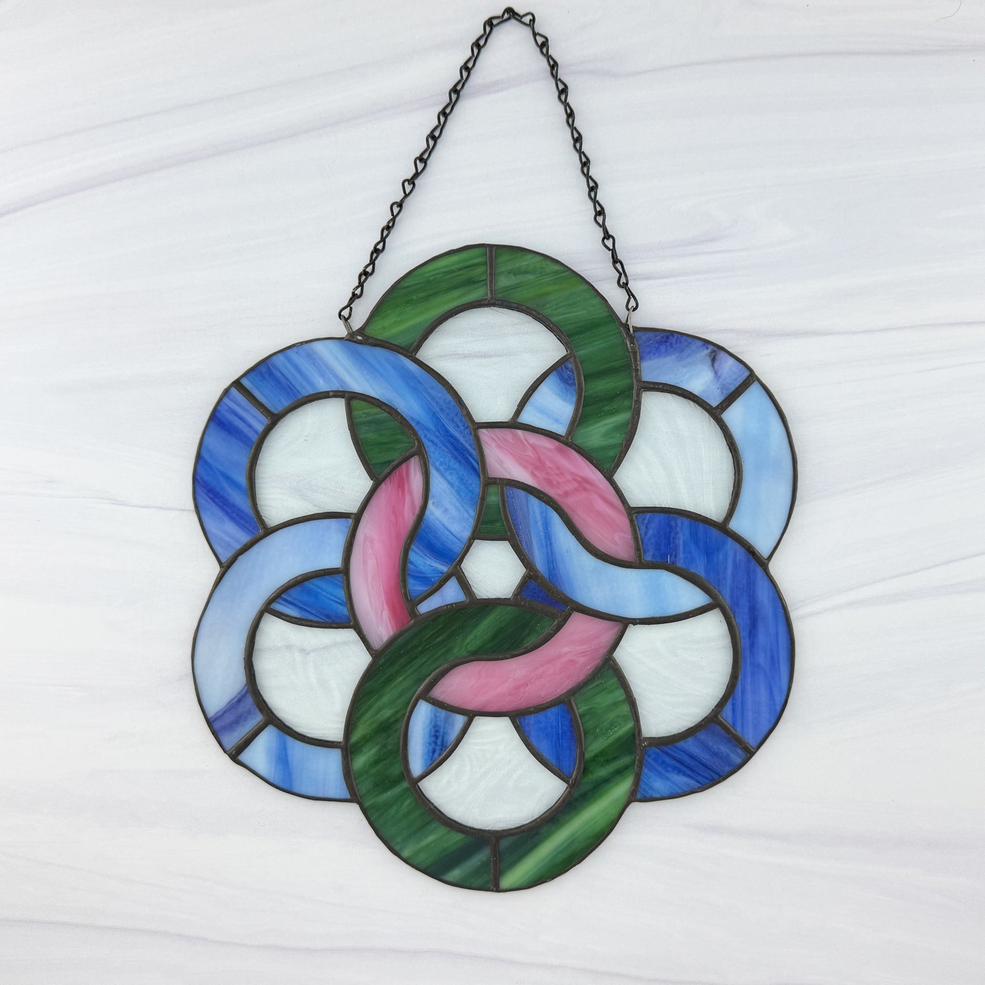 Stained glass window hanging with interlocking rings on a white background. The inner ring is of pink and white wavy glass surrounded by dark blue, light bluish-white and textured green glass rings.