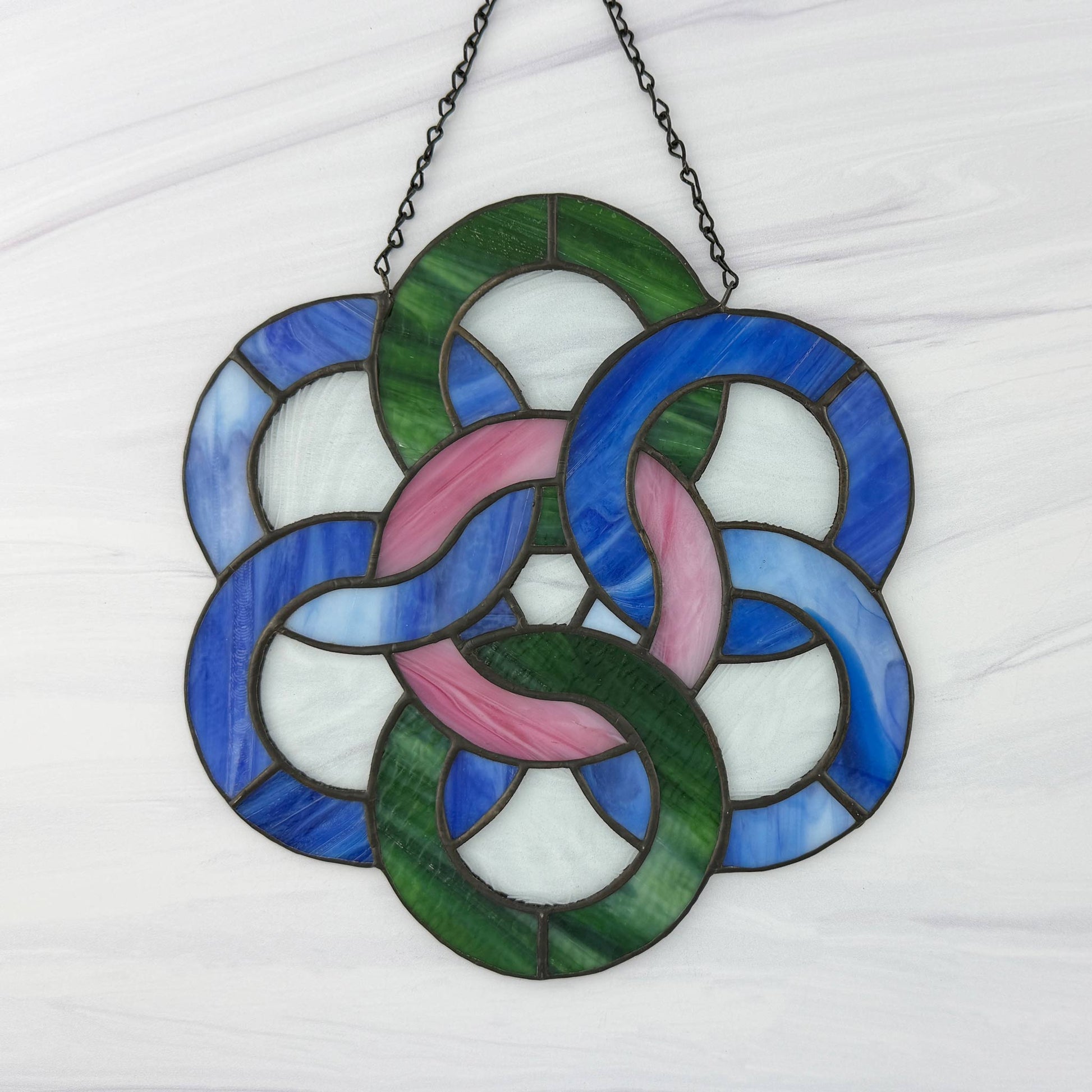 Stained glass window hanging with interlocking rings in blue, green, and pink on a white background.
