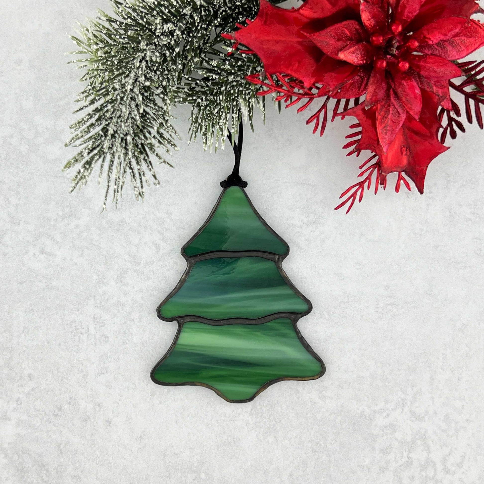 A stained glass multi-colored green Christmas tree ornament. There are swirls of white in the green glass that make it feel like snow is falling on the tree.